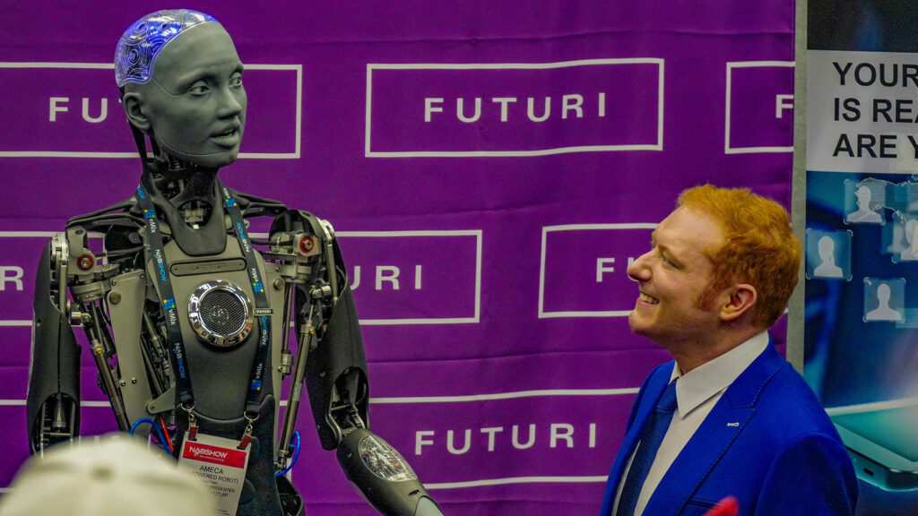 A human-sized robot stands to the left, seemingly looking at a man on the right. A step-and-repeat backdrop reads "Futuri"